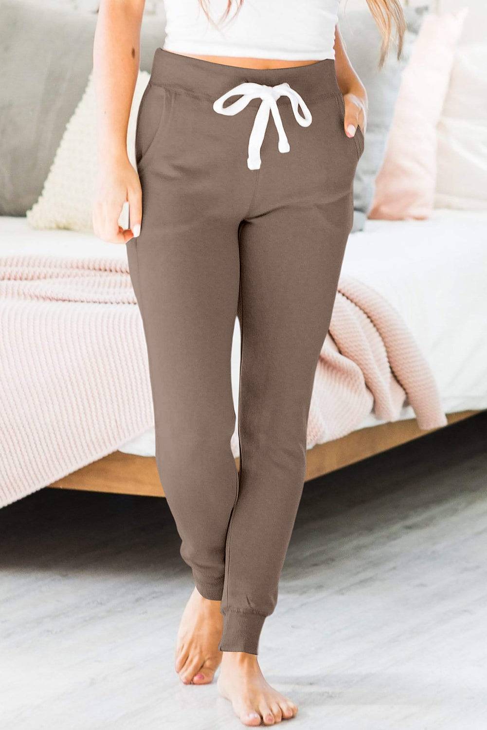 a woman standing in front of a bed wearing brown pants