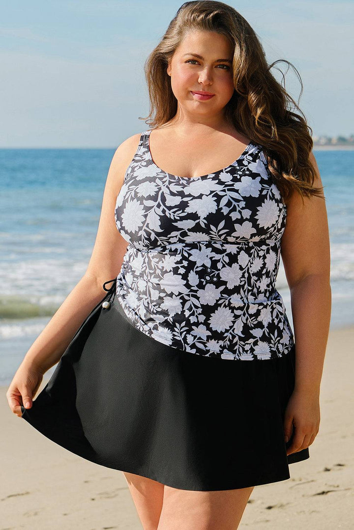 Black Plus Size Printed Square Neck Drawstring Ruffled Tankini Set with floral top and black skirt, modeled on beach. Shop at Faith & Co. Boutique.